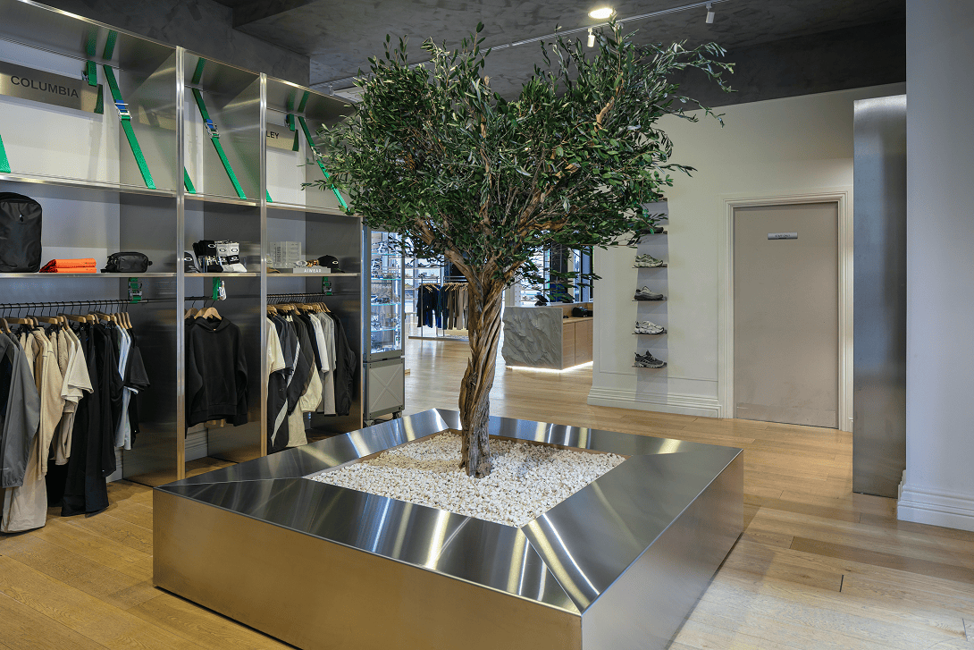 Feature olive tree display within the Drip Moda Liverpool retail store, incorporating bespoke stainless steel-clad shelving, a custom stainless steel tree planter and limewash decorative finishes as part of the premium retail fit-out delivered by Bobblehat.