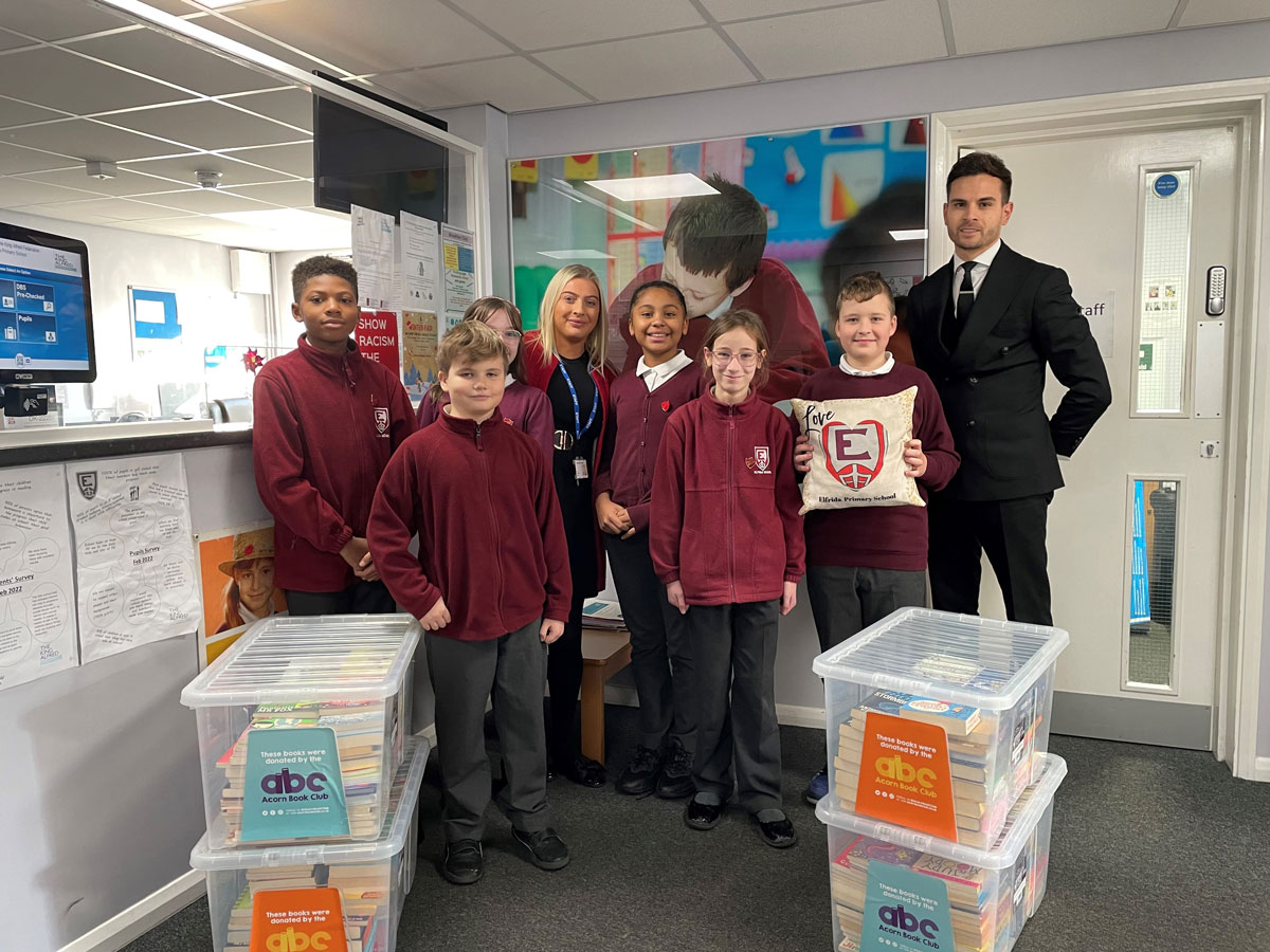 Group of seven schoolchildren in maroon uniforms with two adults standing in an indoor setting, flanked by plastic containers filled with books labeled 'Acorn Book Club'.