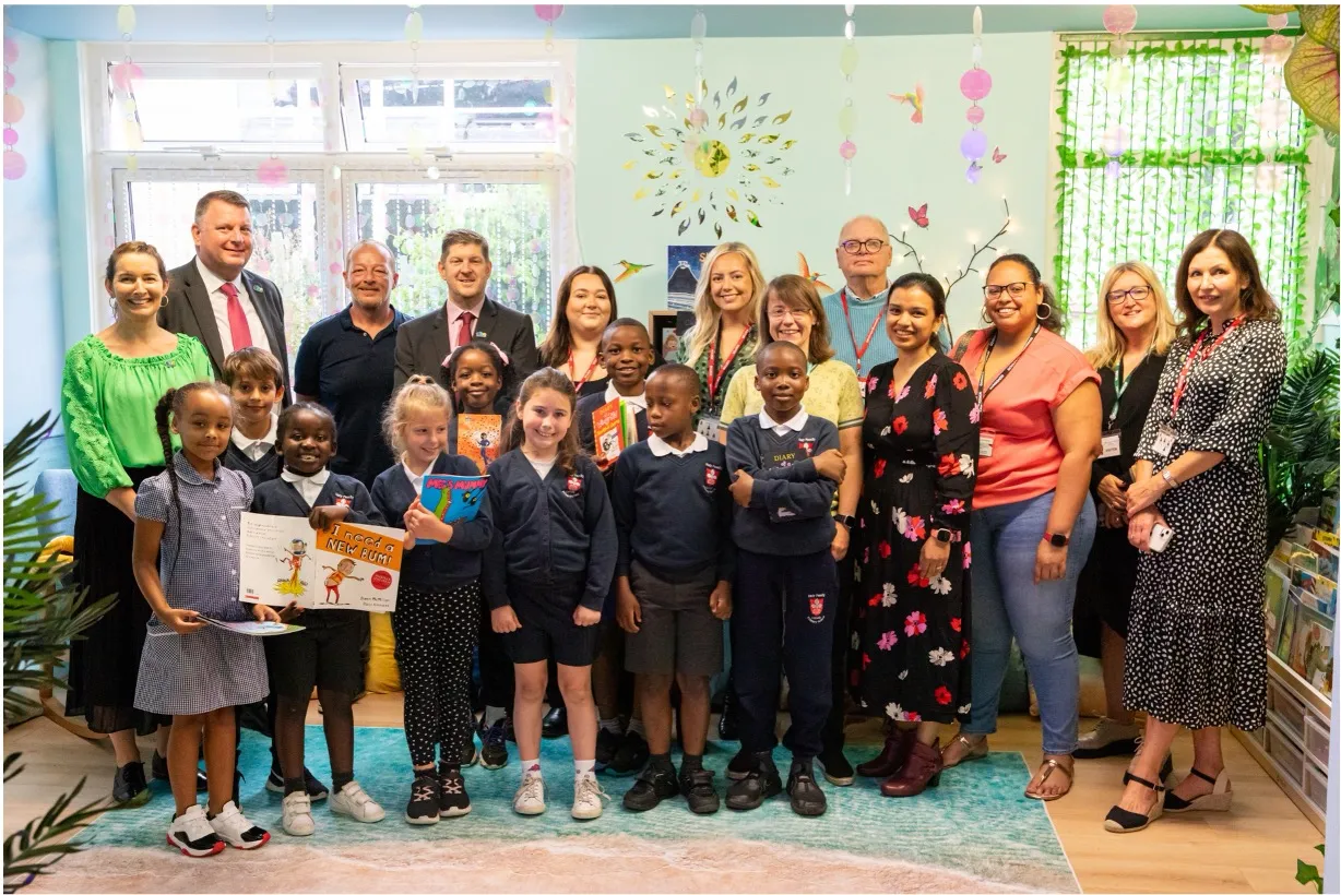 Group photo of diverse school children and adults in a decorated classroom holding books, smiling at the camera.