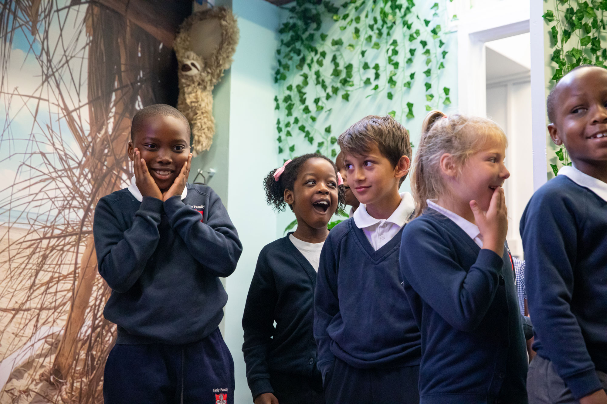 Group of diverse schoolchildren in navy blue uniforms sharing smiles and laughter indoors with green leafy wall decor.