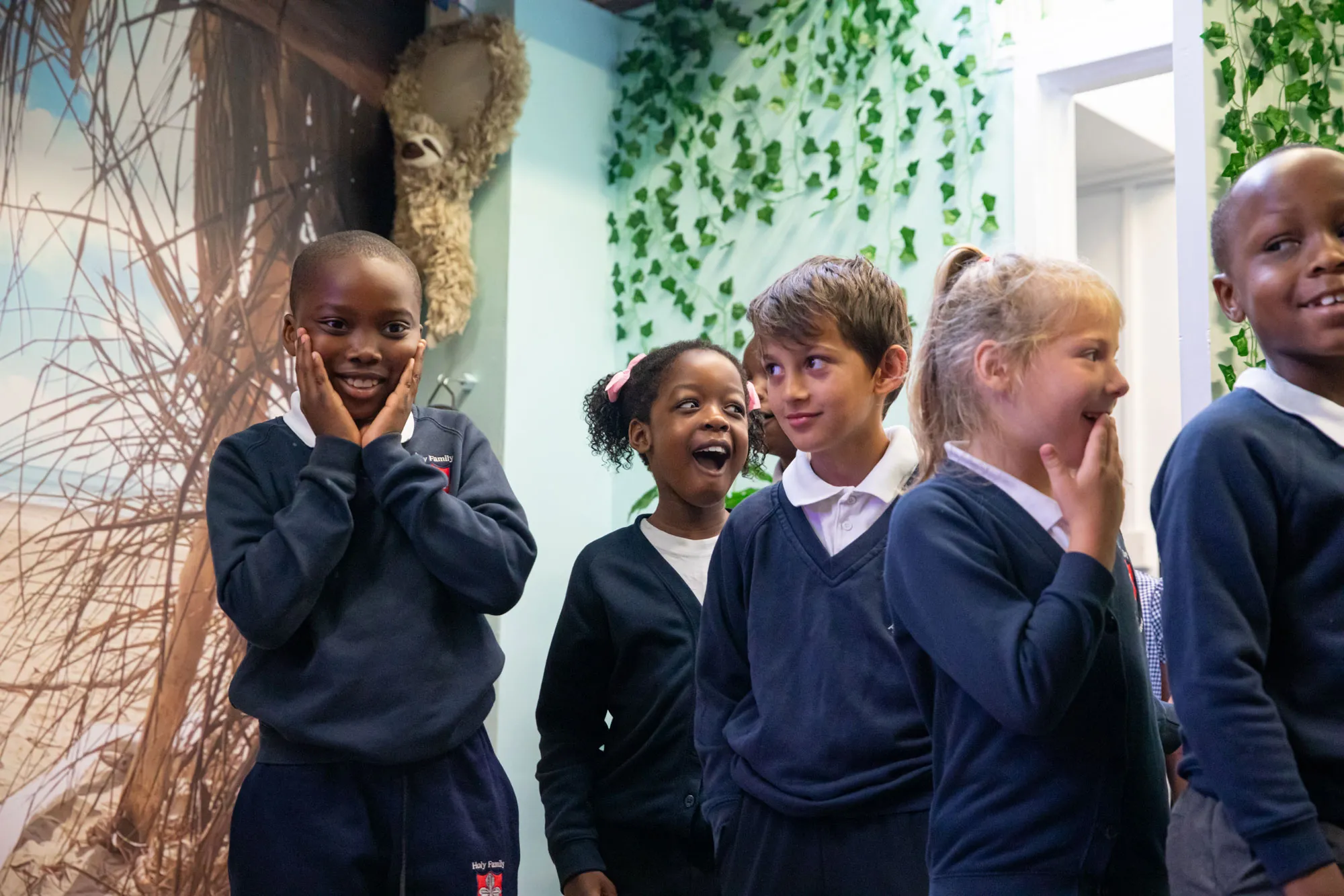 Group of diverse schoolchildren in navy blue uniforms sharing smiles and laughter indoors with green leafy wall decor.
