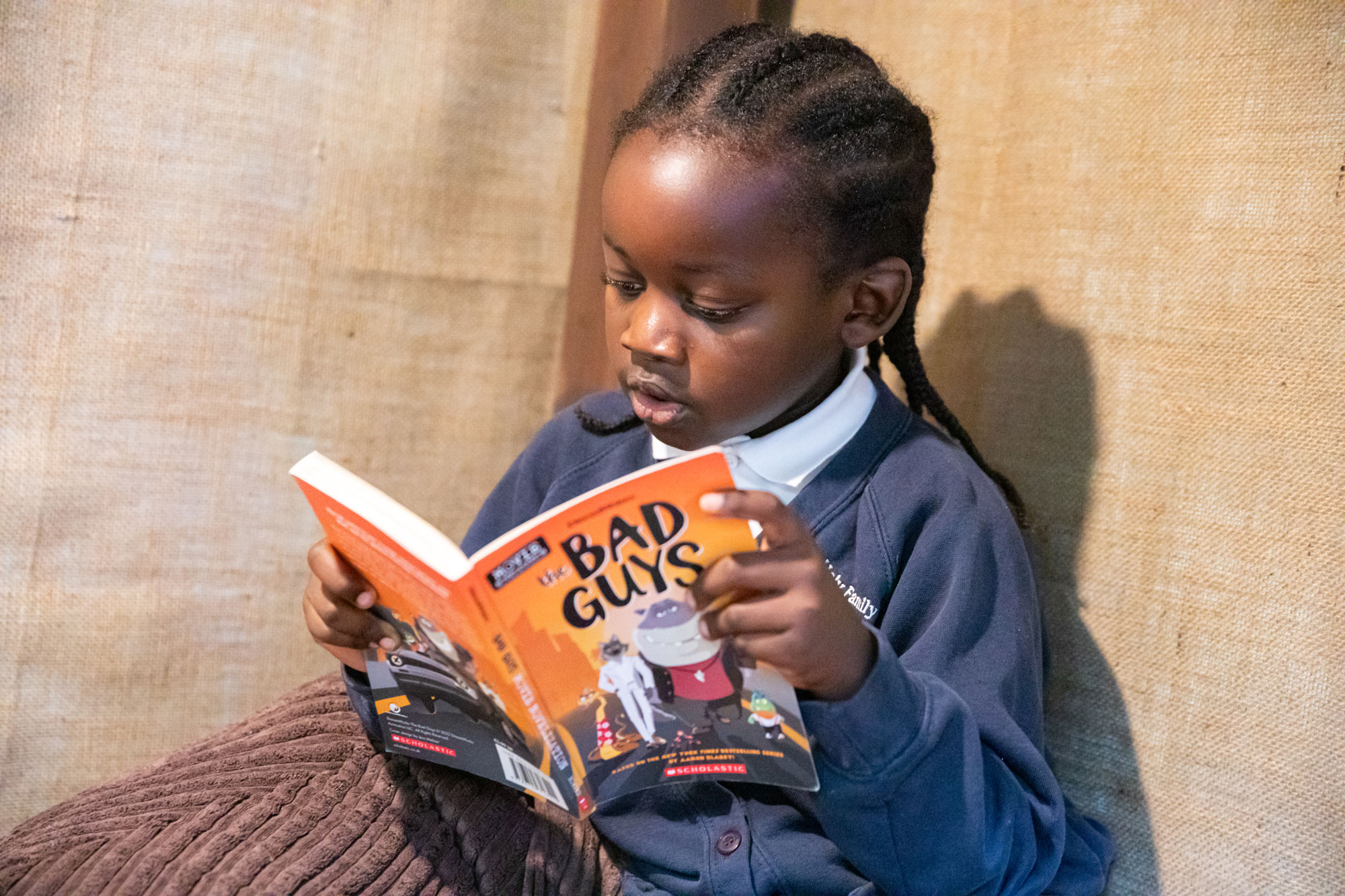 Young child with braided hair reading the book 'The Bad Guys' while sitting on a brown cushion.