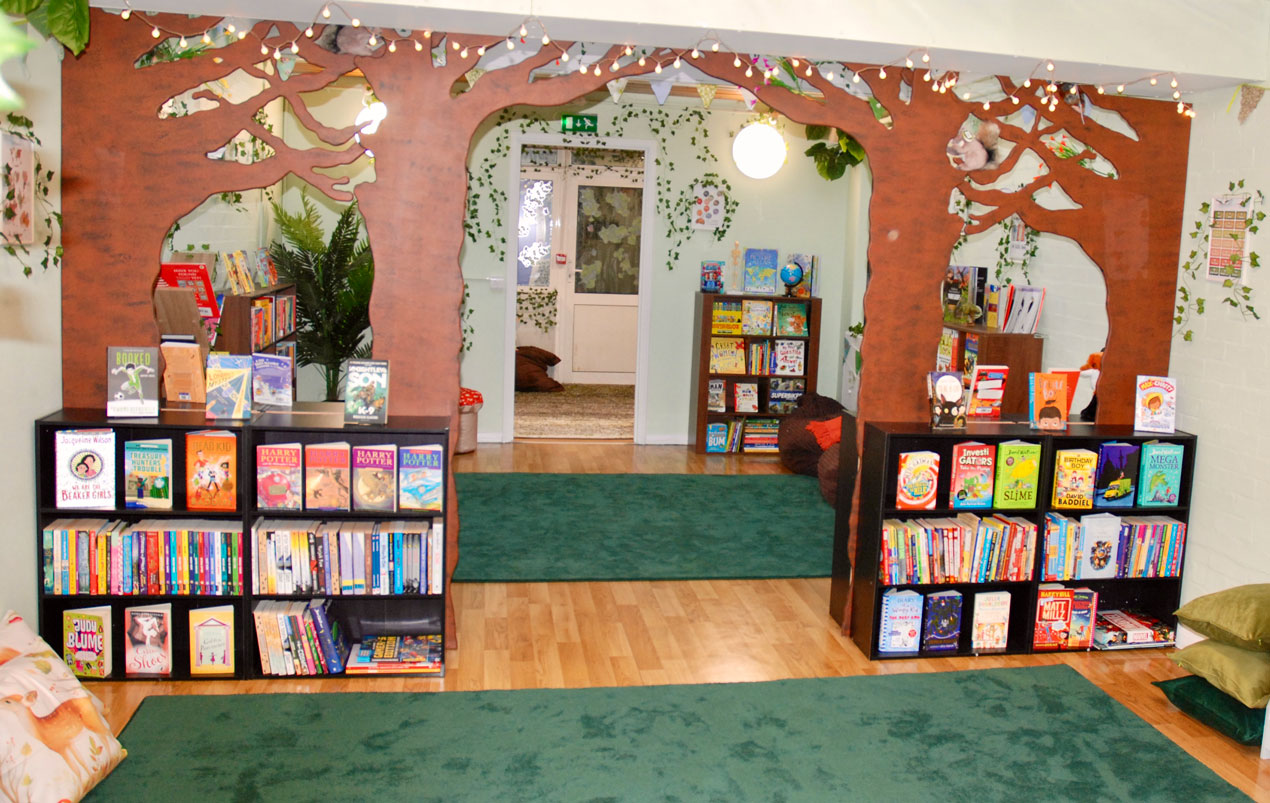 Children's reading area with bookshelves filled with colorful books, tree-shaped wooden archway with hanging lights, green rugs, and cozy seating cushions.