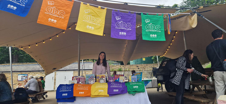 Outdoor bookstall under canopy with colorful abc book bags hanging and displayed, a woman standing behind the table filled with books, and people seated at picnic tables nearby.