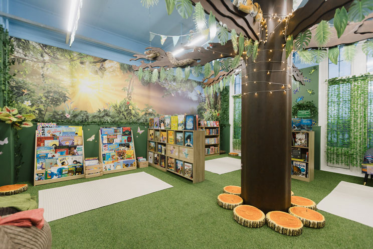 Children's reading area with artificial grass, bookshelves filled with colorful books, a large artificial tree wrapped in string lights, and tree stump cushions arranged around its base.