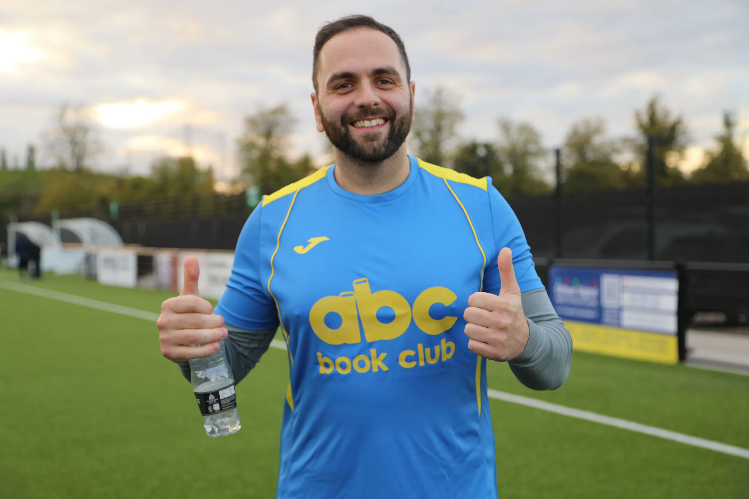 Smiling man in blue and yellow sports jersey holding a water bottle and giving thumbs up on a soccer field.