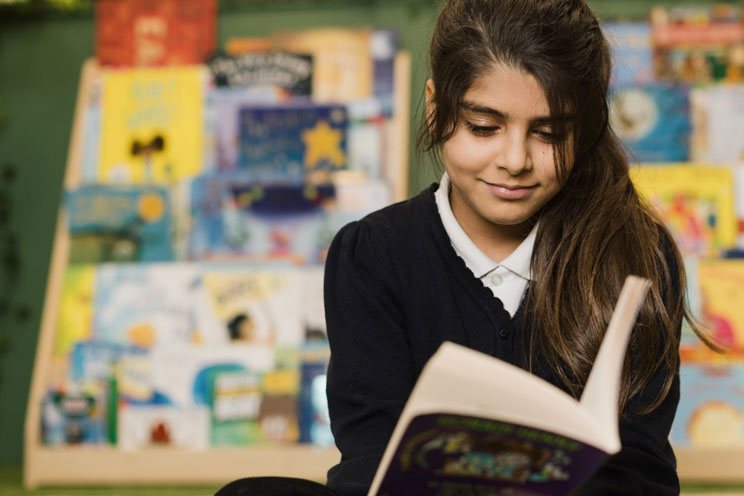 Young girl with long brown hair reading a book in front of a blurred book display.