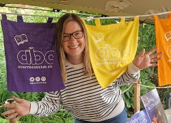 Smiling woman wearing glasses and a striped shirt poses playfully holding a yellow abc scarf with colorful abc scarves hanging in the background.