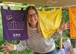 Smiling woman wearing glasses and a striped shirt poses playfully holding a yellow abc scarf with colorful abc scarves hanging in the background.