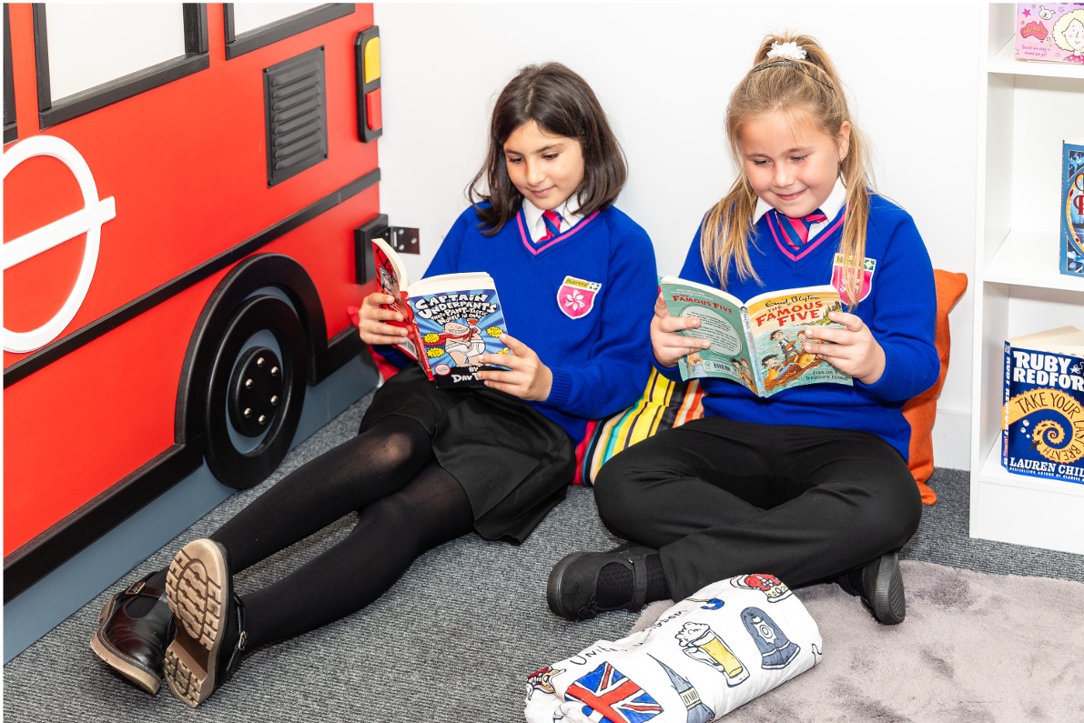 Two schoolgirls in blue sweaters and black skirts sitting on the floor reading books in a classroom corner with a red bus decoration nearby.