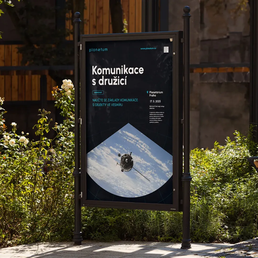 Poster for Planetum Praha event “Communication with satellite”, placed in an outdoor advertising stand among the greenery.