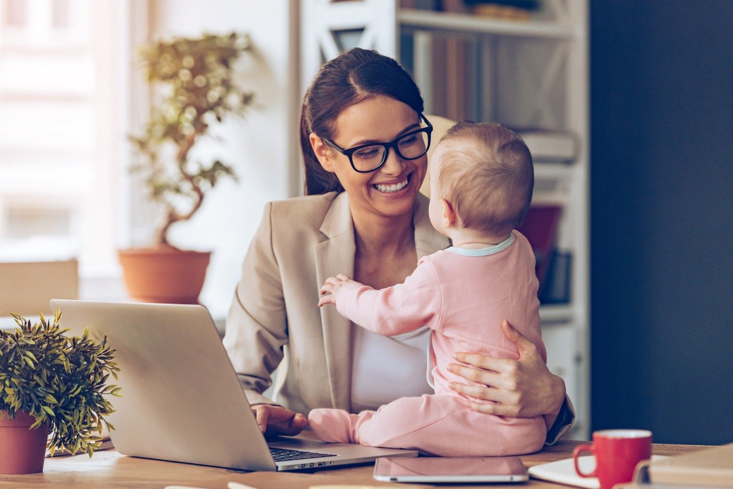 Working mother holding baby while focusing on laptop in home office