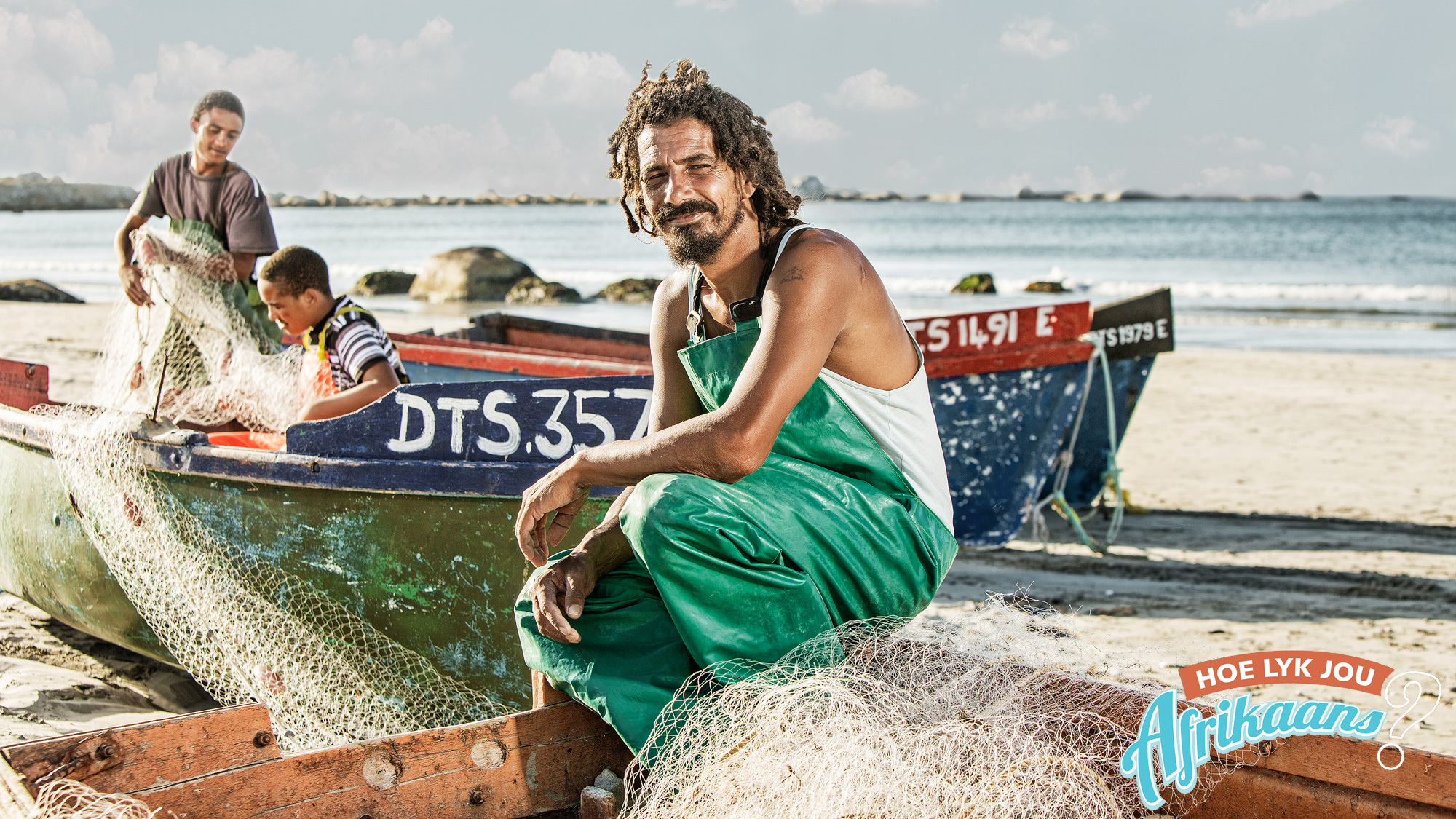 Smiling fisherman in green overalls sitting on a boat with fishing nets on a sandy beach, with two others handling nets on boats in the background.