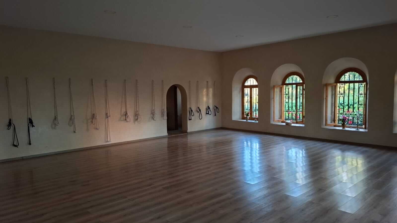 Empty yoga or exercise studio with wooden floors, beige walls, ropes hanging on one wall, and arched windows with bars and flower pots.