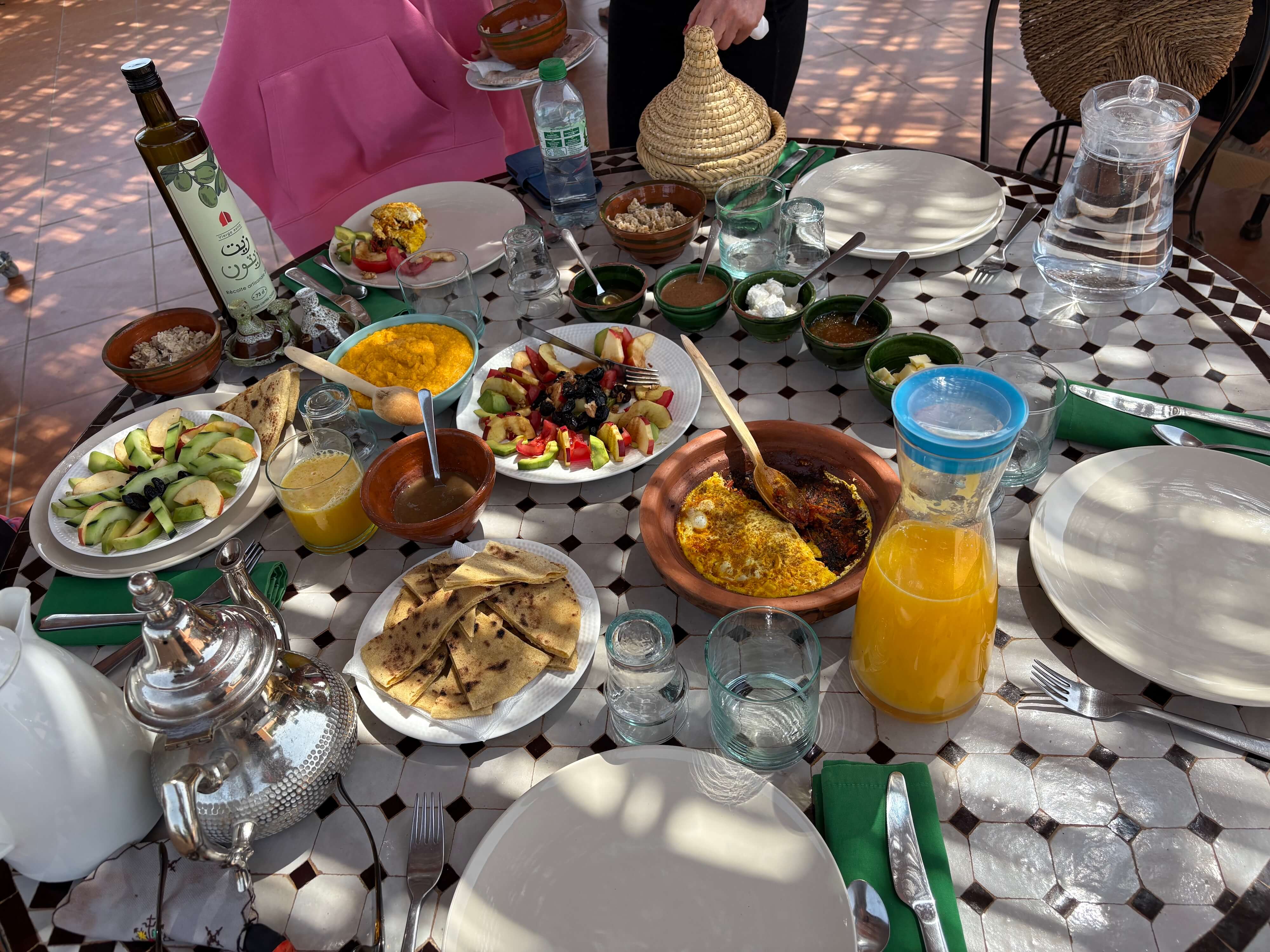 Table set with Moroccan-style dishes including flatbread, fruit salad, orange juice, sauces, and a teapot under dappled sunlight.