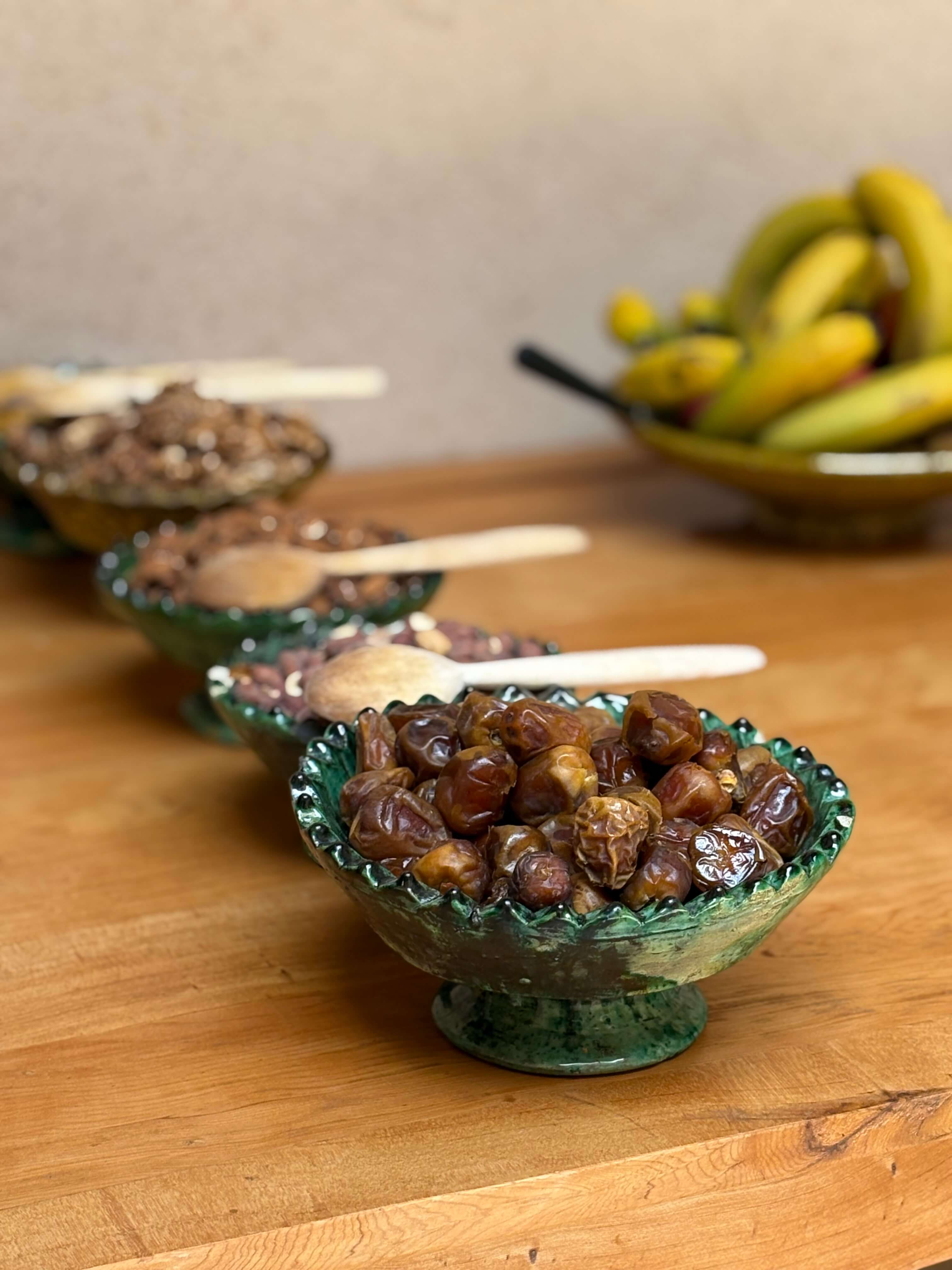 Green ceramic bowl filled with dried dates in the foreground, with other bowls containing nuts and a bunch of bananas in the background on a wooden table.