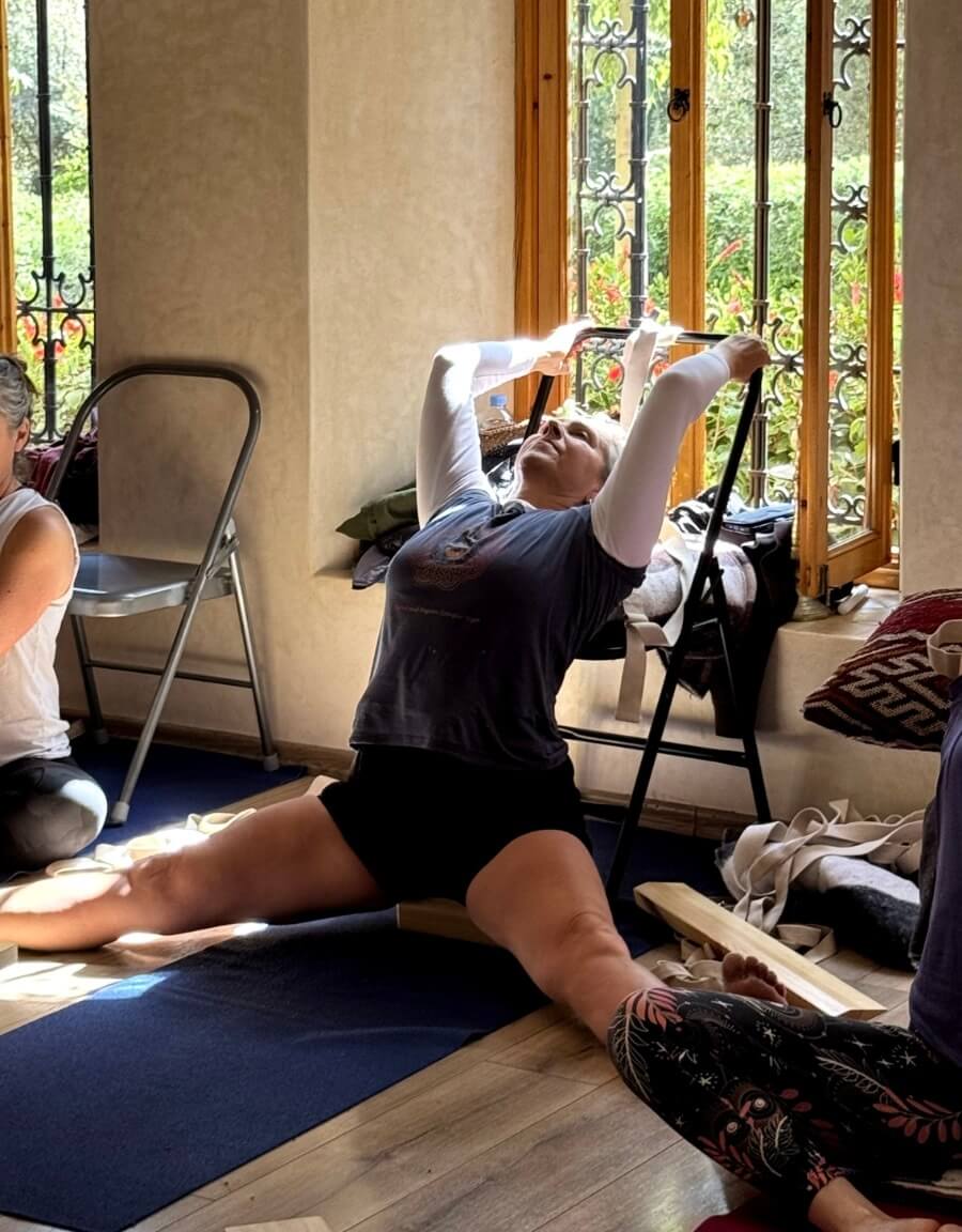 Woman doing a backbend stretch using a chair for support in a sunlit room during a yoga session.