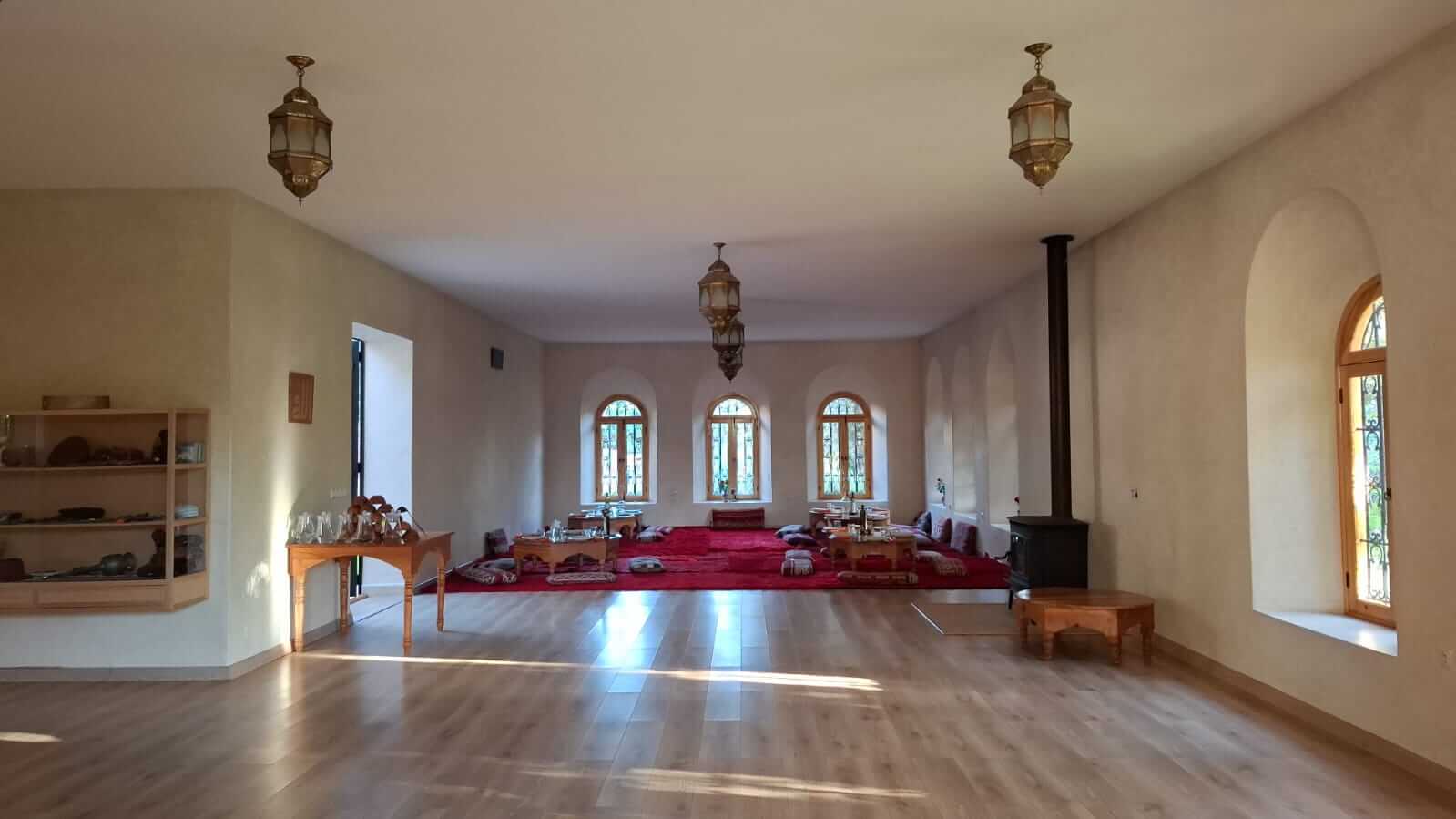 Spacious room with wooden floor, arched windows, hanging lanterns, and a low seating area with red carpet and cushions.