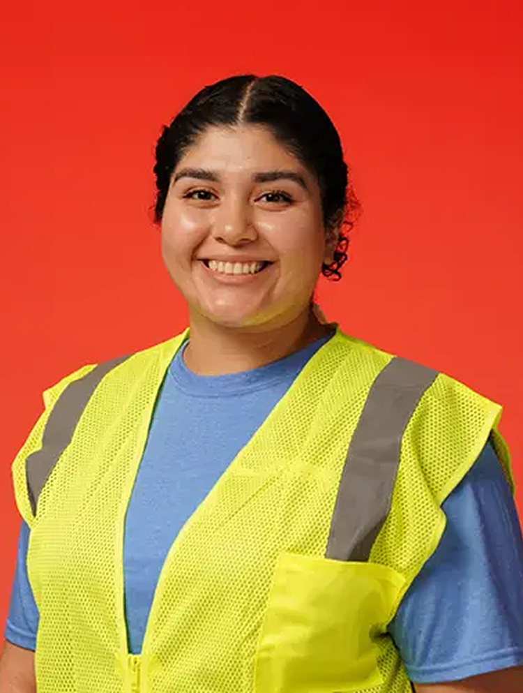 Smiling person with dark hair wearing a blue shirt and yellow safety vest against an orange background.