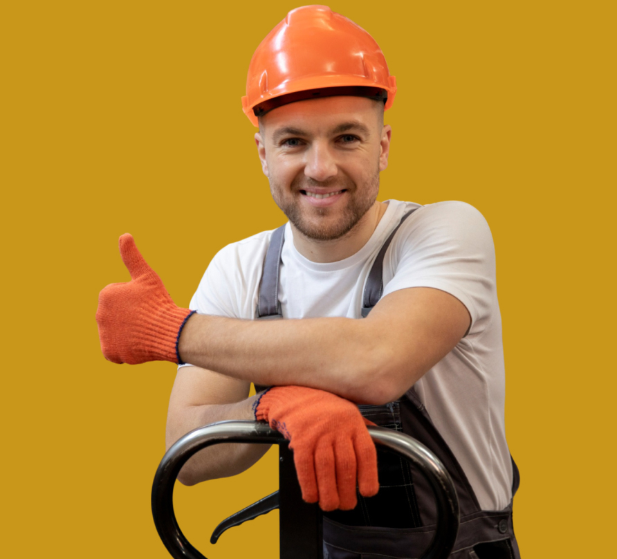 Smiling construction worker wearing an orange hard hat and gloves, giving thumbs up while leaning on equipment.