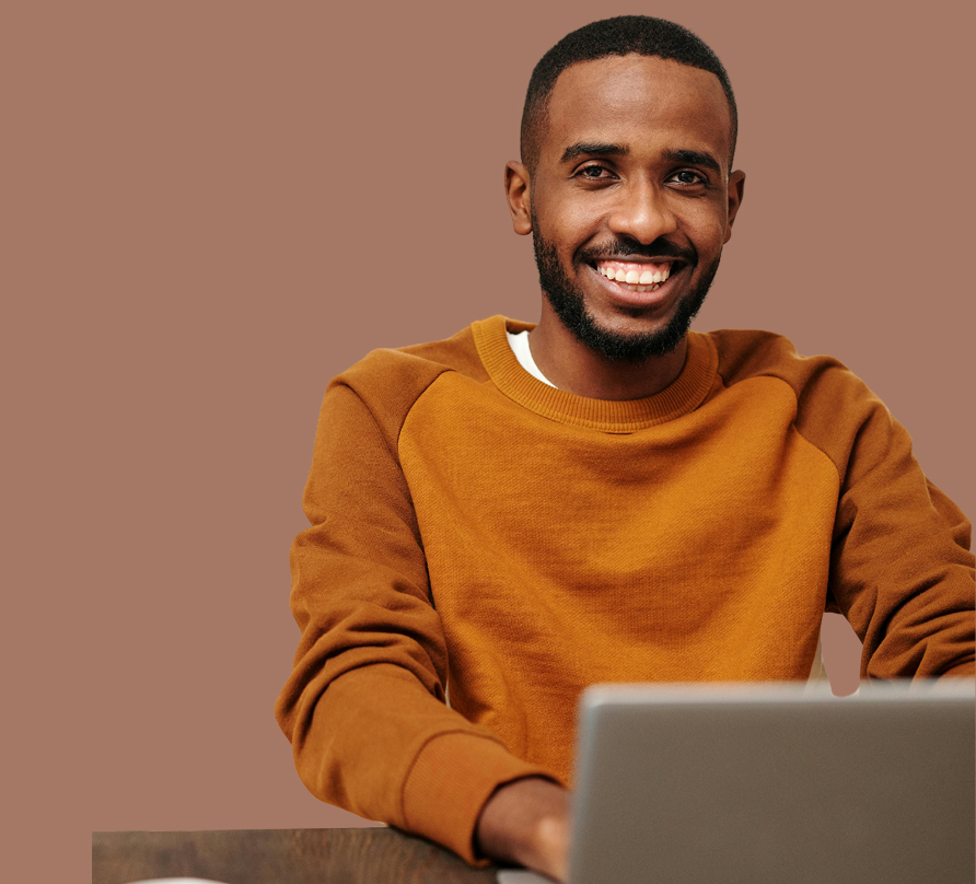 Smiling man in brown sweater working on a laptop against a purple background.