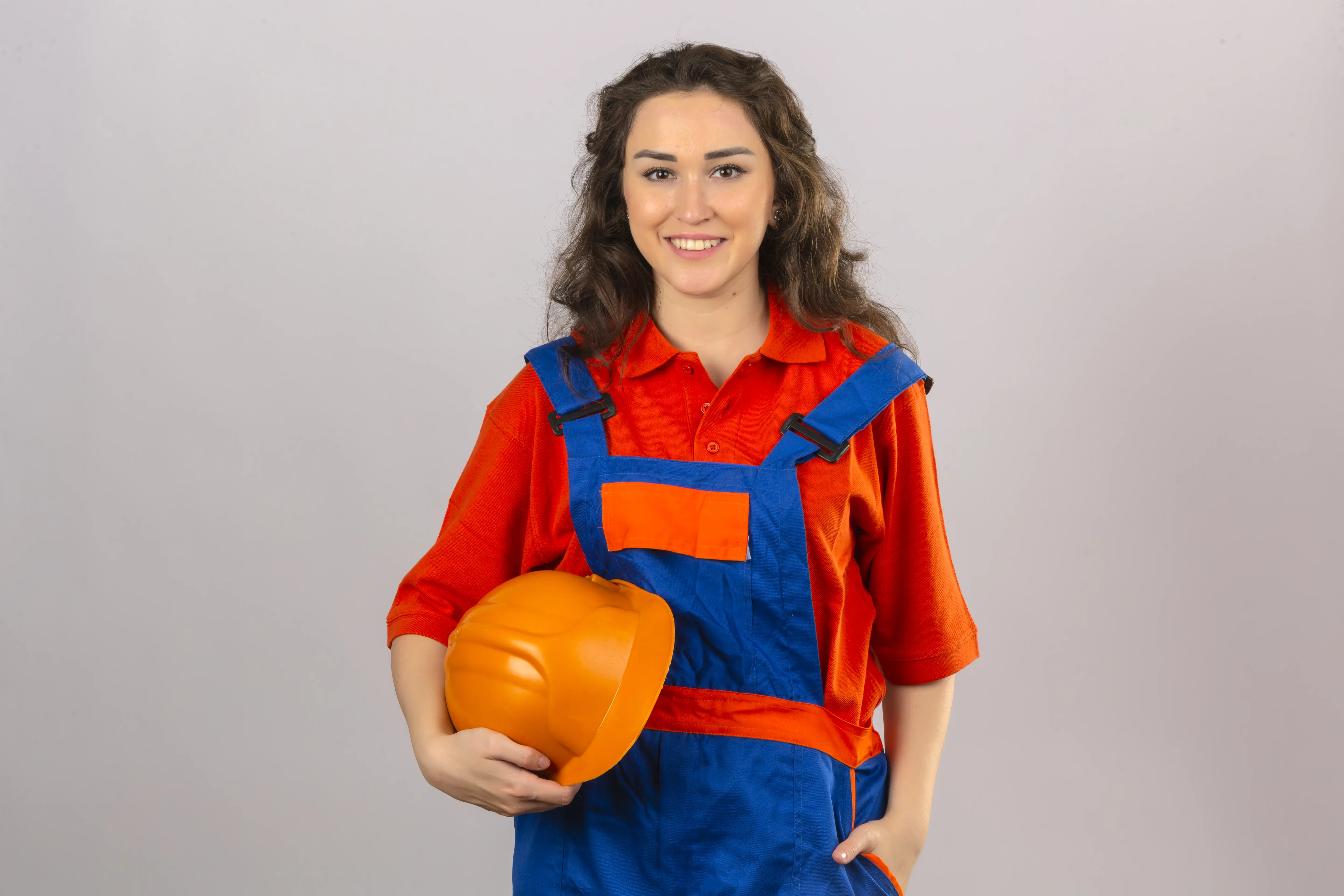 Smiling young woman wearing orange and blue work overalls holding an orange safety helmet.