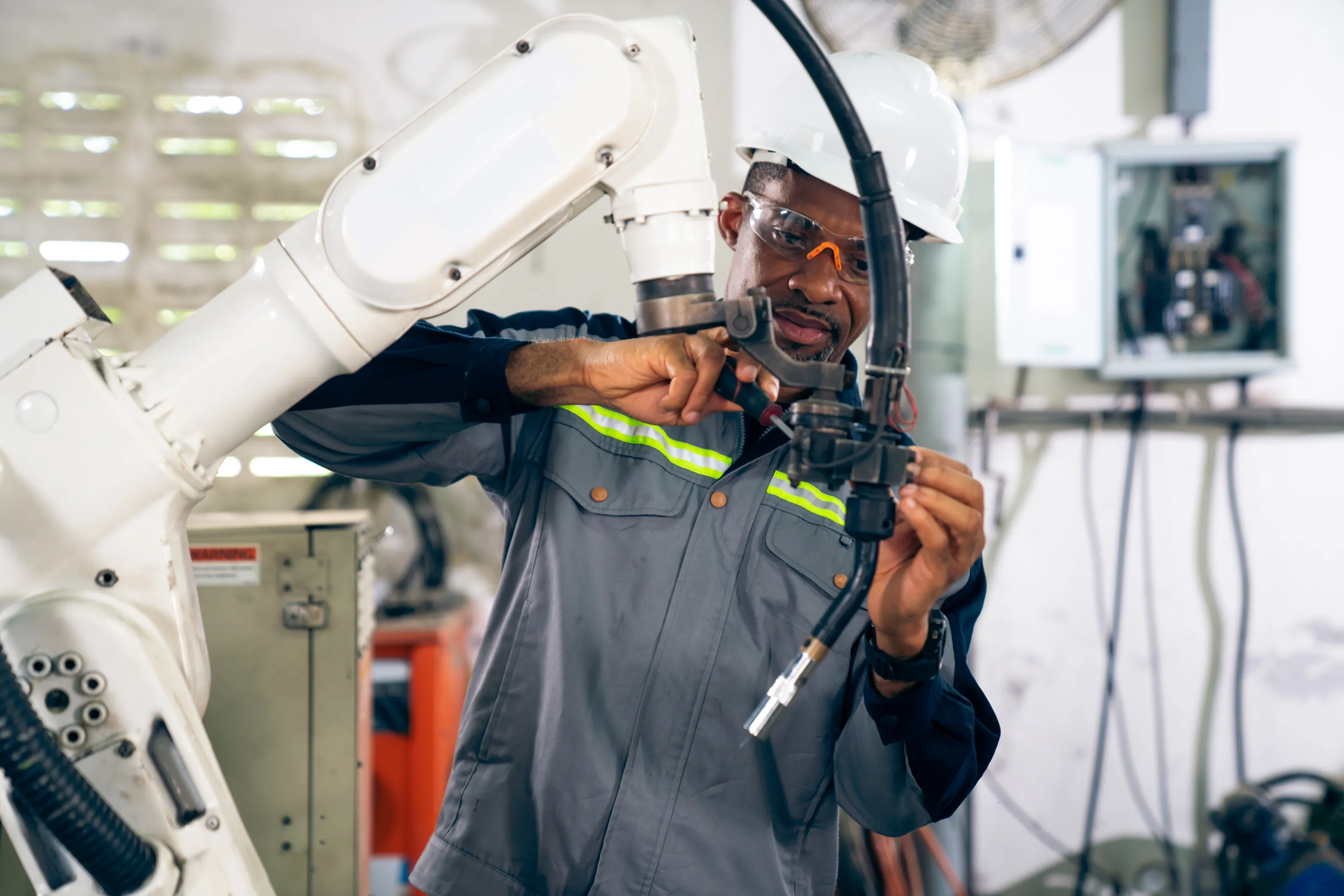 African American man wearing safety glasses and hard hat, repairing robotic arm using screwdriver.