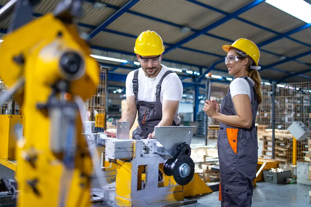Two factory workers in protective gear operate machinery in an industrial setting, surrounded by equipment and metal structures.