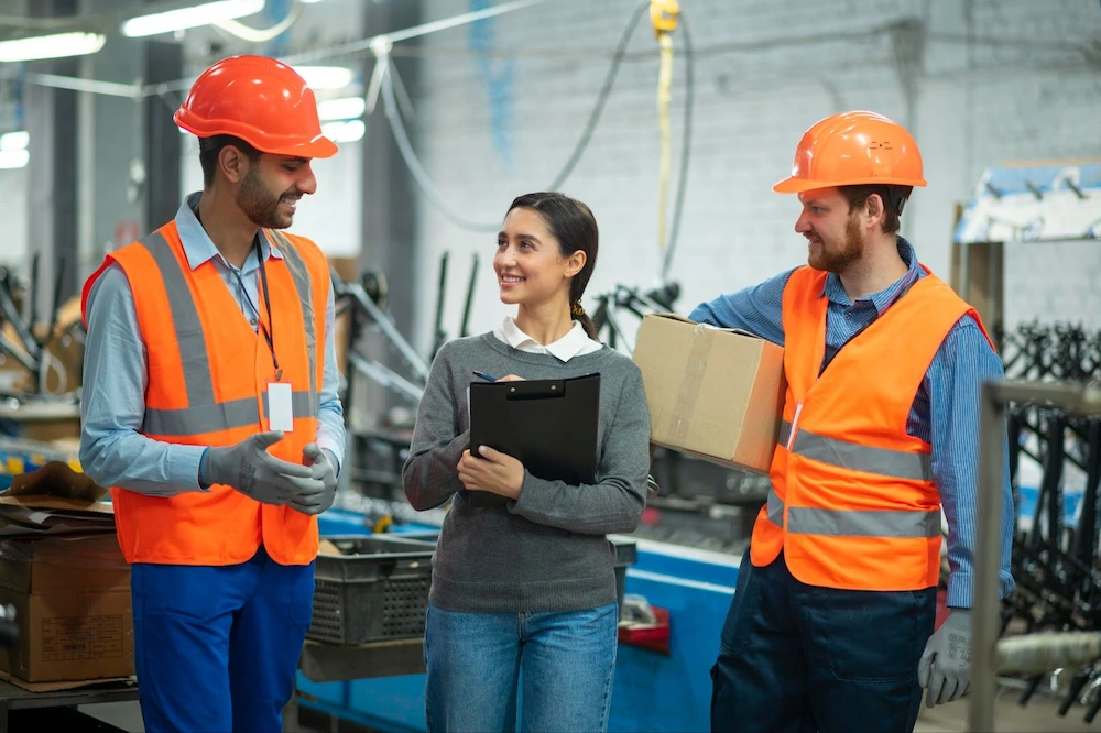 Two workers in safety vests and hard hats discuss logistics while a woman holds a clipboard in a busy industrial setting.