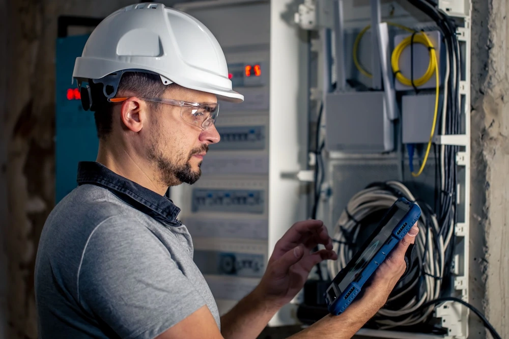 An individual in a hard hat examines a tablet near a wall-mounted electrical panel filled with wires and circuit labels.