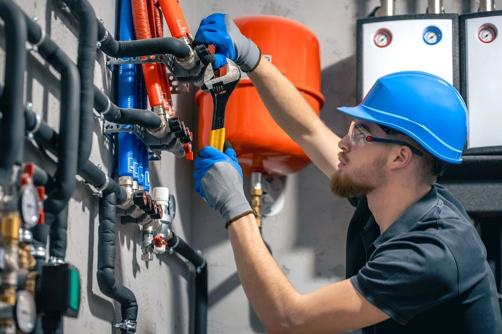 A worker in a blue hard hat uses a wrench to adjust black pipes in an industrial setting, surrounded by gauges and valves.