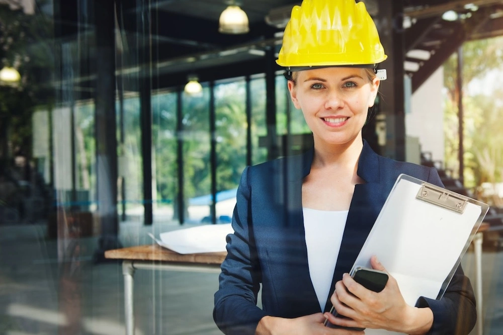 Woman wearing a yellow hard hat and blazer holds a clipboard and smartphone, standing in a modern office with large windows.