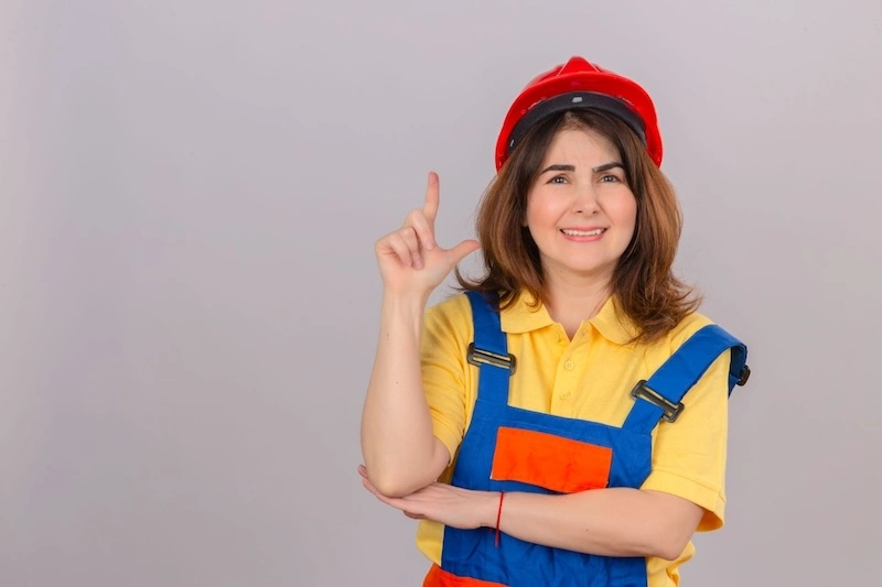 A woman in blue overalls and a red hat points upward with a smile, standing against a bright outdoor background.