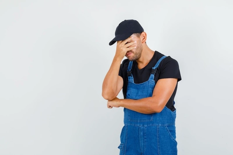 A man in overalls stands against a blue wall, covering his face with his hand.