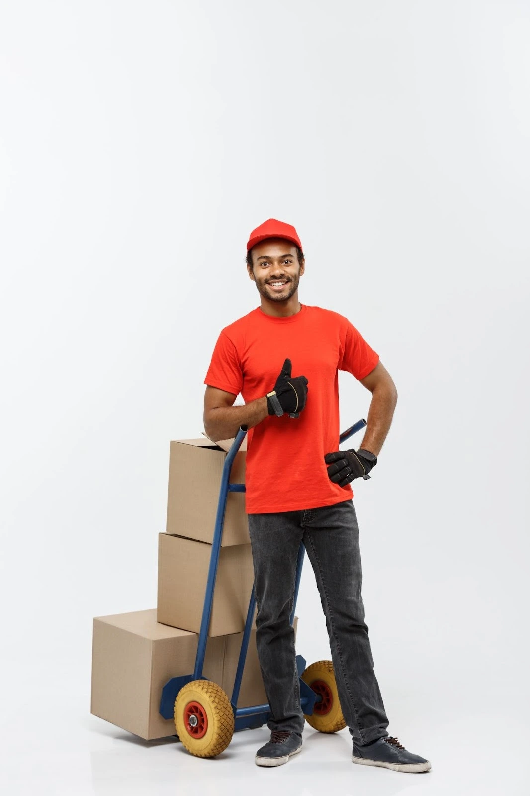 Smiling delivery worker in a red shirt and cap gives a thumbs up, standing beside a blue hand truck with stacked boxes. Positive and energetic mood.