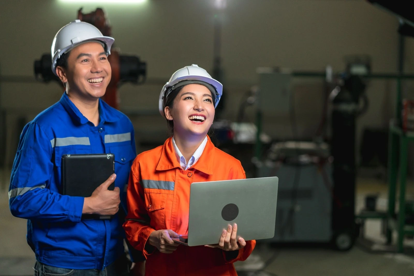 Two engineers in safety helmets, one in blue holding a tablet and the other in orange with a laptop, laughing in an industrial setting.