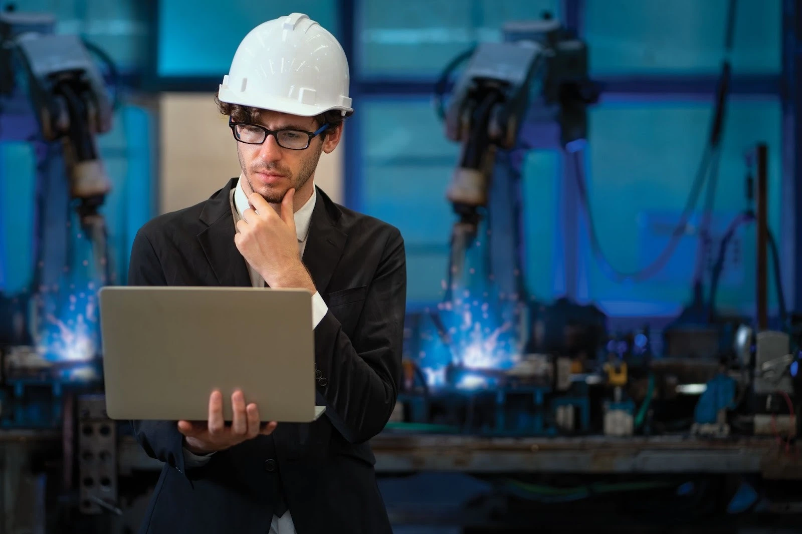 A man in a suit and white hard hat studies a laptop thoughtfully in a factory setting, with robotic arms and sparks behind him, conveying focus and technology.
