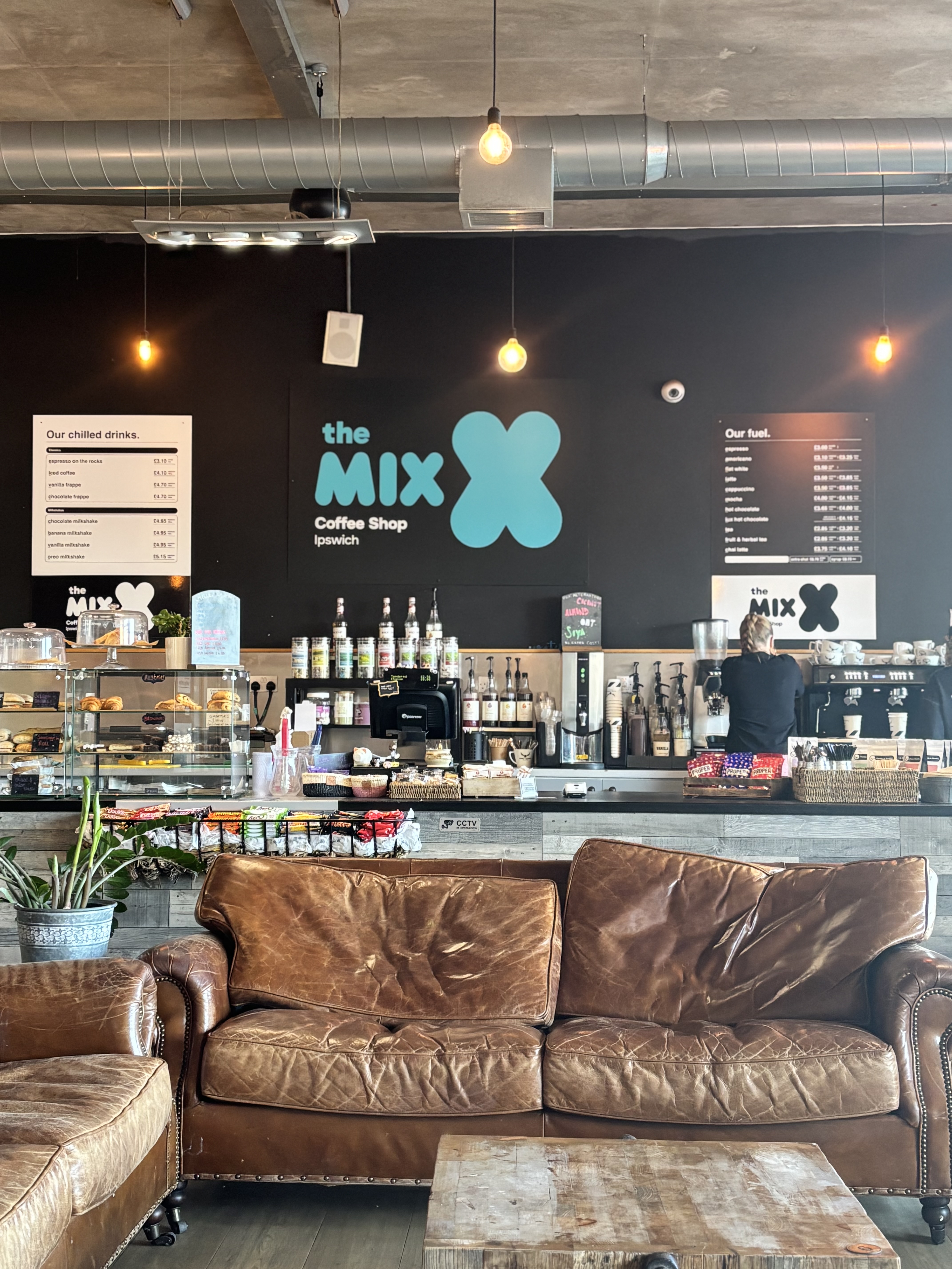 The image shows brown leather chairs in the front with the coffee bar behind with The Mix Coffee Shop branding in teal on the black wall at the new site on Ipswich waterfront in the University of Suffolk building.