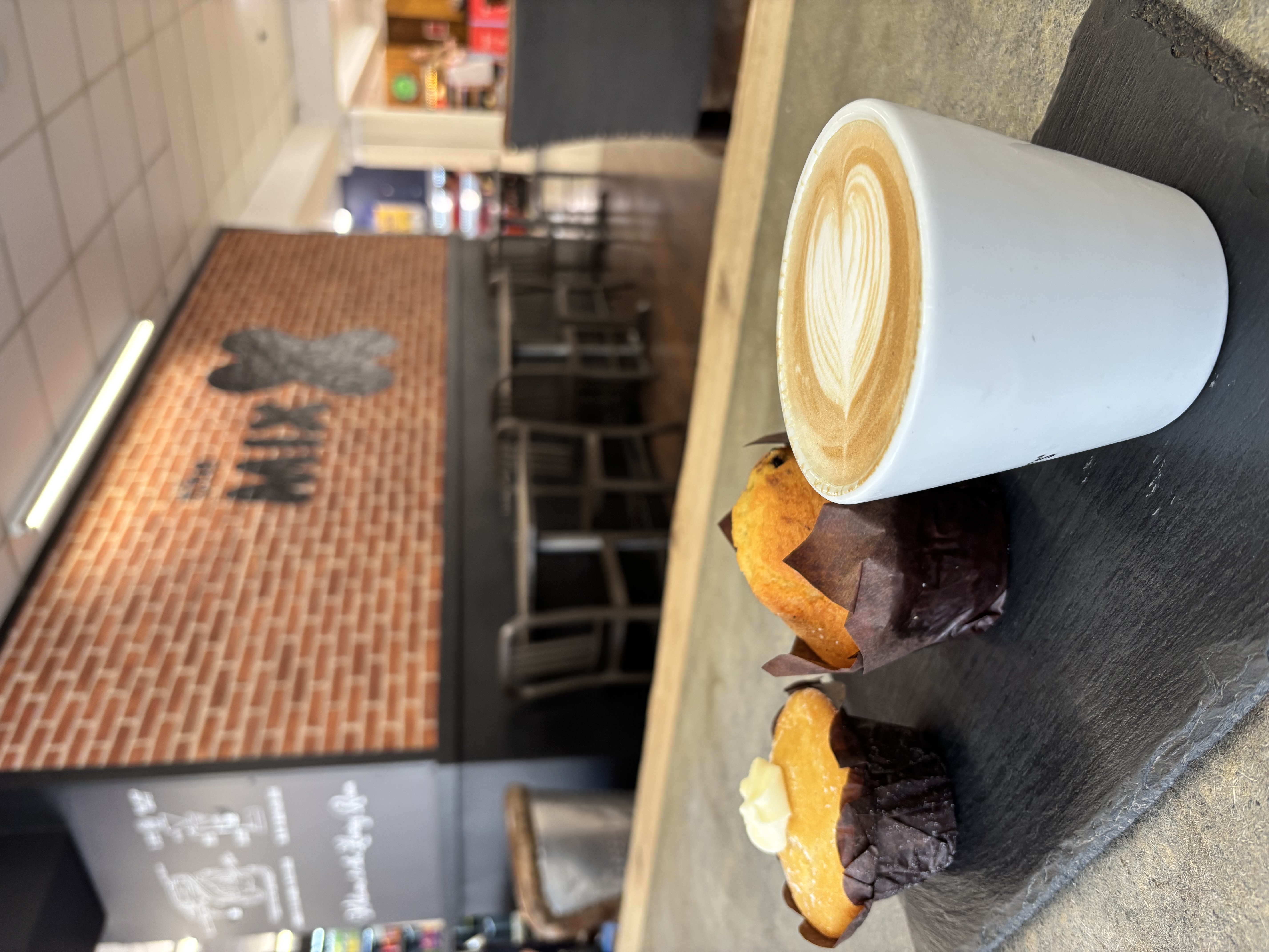 The image shoes a coffee and two muffins on a slate on a table in the foreground, with an exposed brick wall with The Mix logo in black on the wall, inside the new The Mix Coffe Shop Hadleigh which is inside the Co-op on Hadleigh High Street.