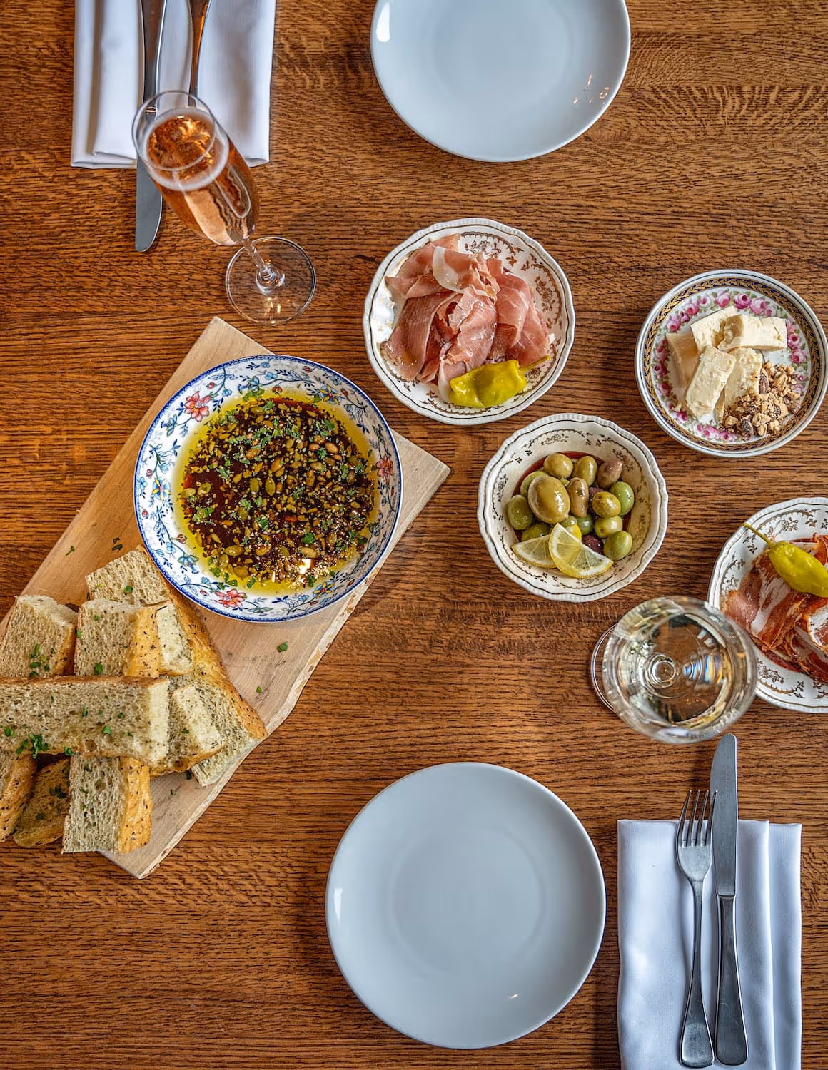 A variety of cheese, olives and cold cuts plated on a clean wood table, set with glasses of wine, focaccia bread and cutlery.