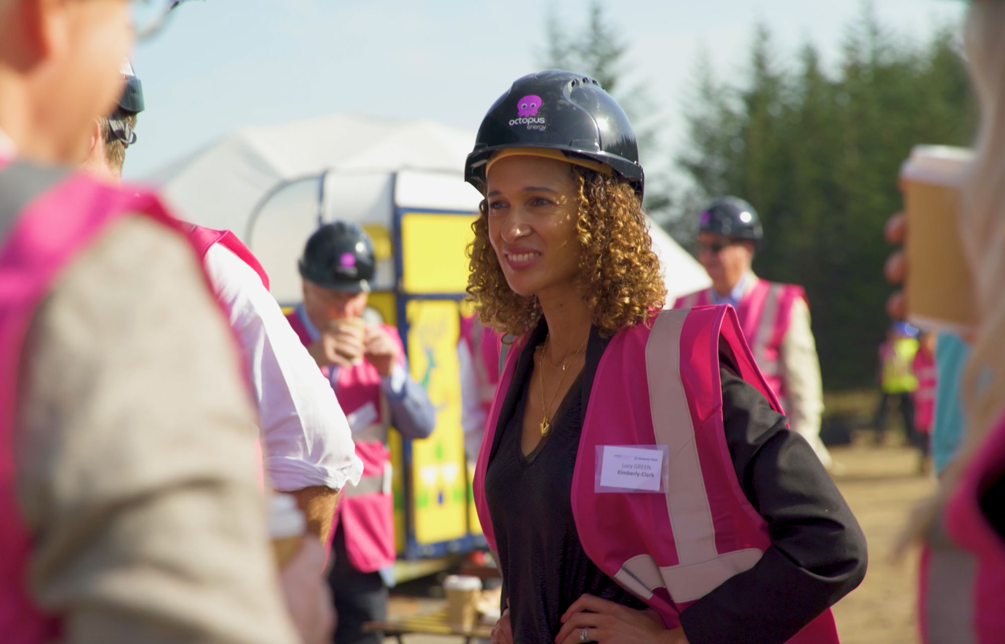 A woman wearing a hard hat and pink safety vest participates in an outdoor construction event.