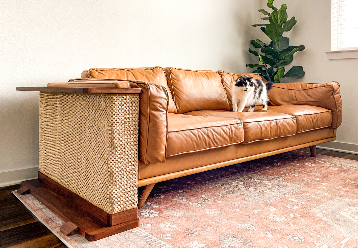 A cute black and white cat walking across a leather sofa with a Cat-e-Corner cat scratcher on the corner of the couch.