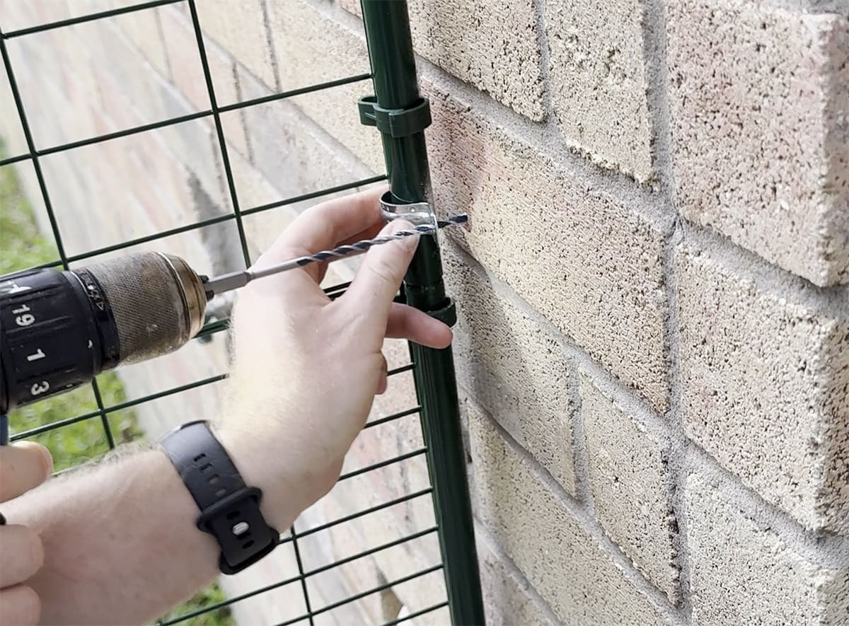 A person's hand holding a drill and a U-bracket who is drilling in to the brick with a masonary bit.