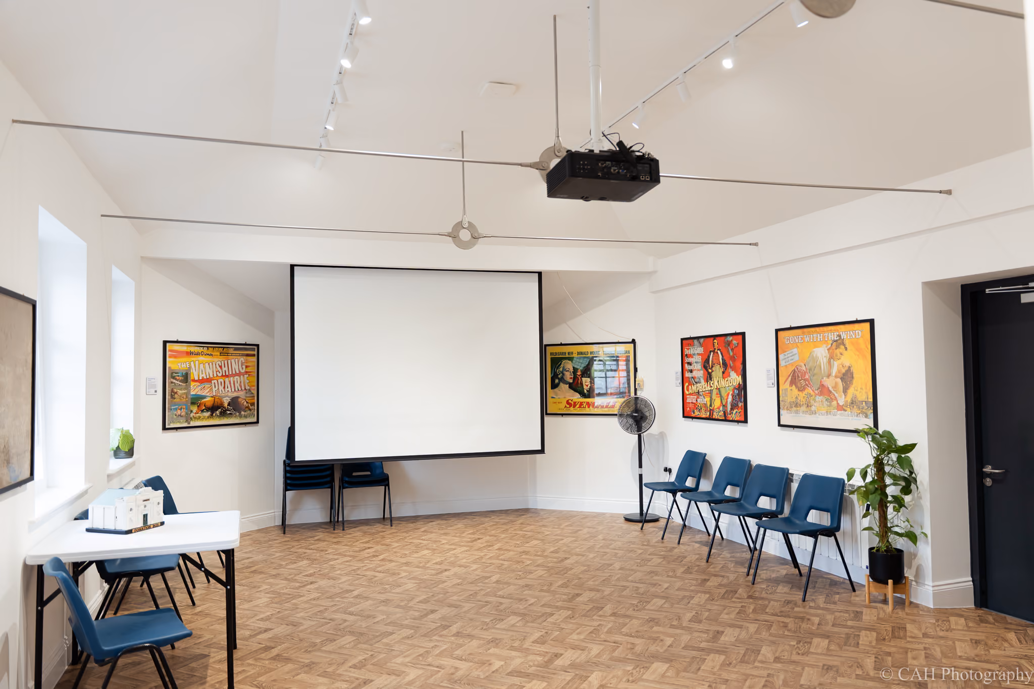 The Pavilion Studio with a large projection screen. Blue chairs line the bright white room with parquet flooring and framed vintage film posters on the walls. ‍Photo credit: CAH Photography.