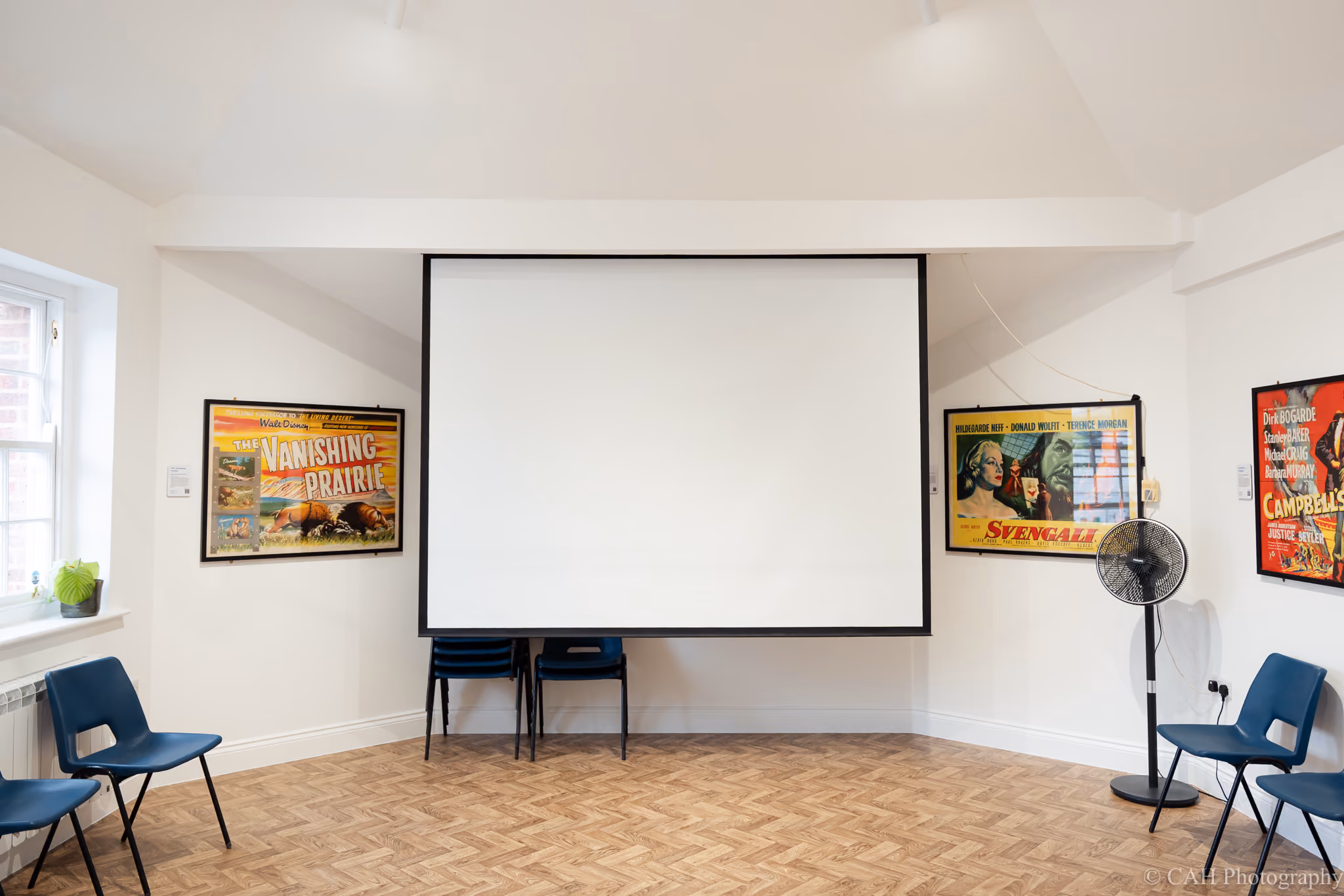 The Pavilion Studio arranged with a large projection screen and blue chairs around a bright room with parquet flooring. Framed vintage film posters include "The Vanishing Prairie" and "Svengali" with part of a "Campbells Kingdom" poster visible beside a standing fan. ‍Photo credit: CAH Photography.