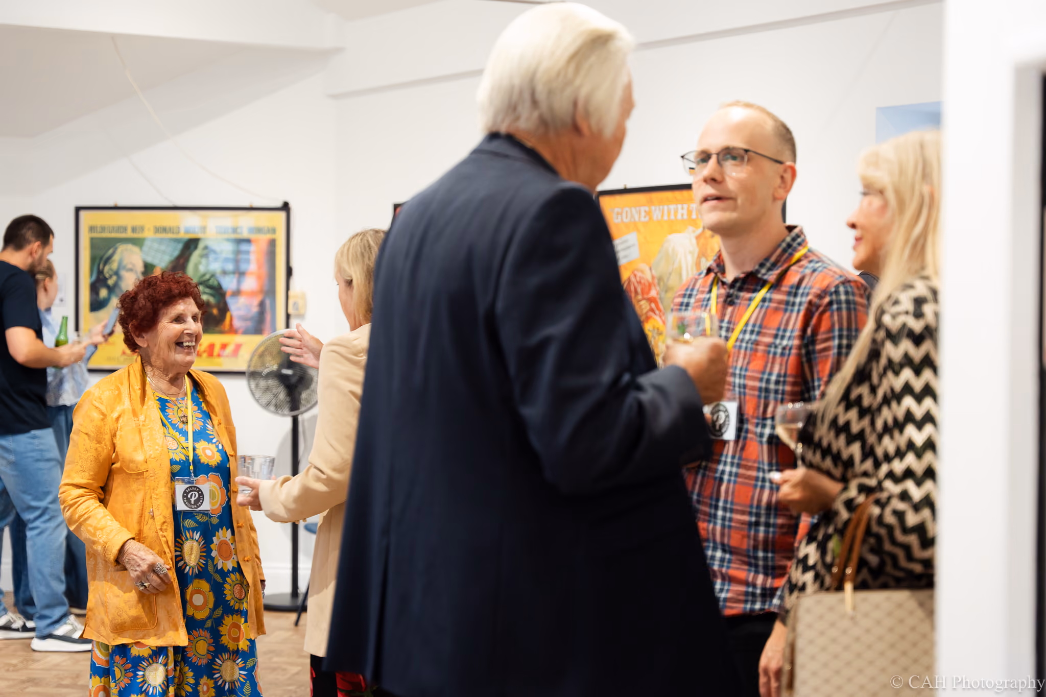 Guests mingle and chat during an event at The Pavilion Studio. An older woman in a bright orange jacket and patterned dress smiles while groups nearby hold drinks and talk beneath vintage film posters including "Gone with the Wind". ‍Photo credit: CAH Photography