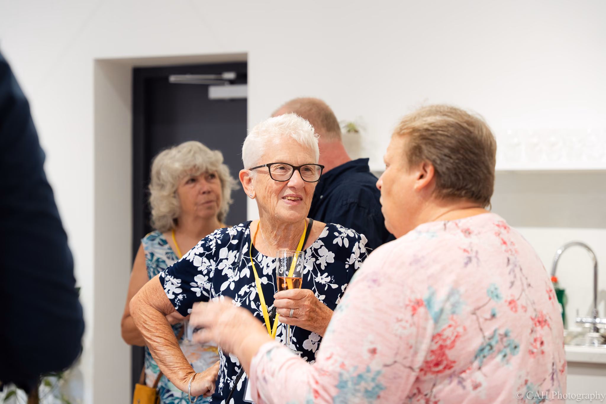 Guests chat and socialize inside The Pavilion Studio during an event. An older woman with short white hair and glasses holds a drink while talking with another guest in the bright white space. ‍Photo credit: CAH Photography