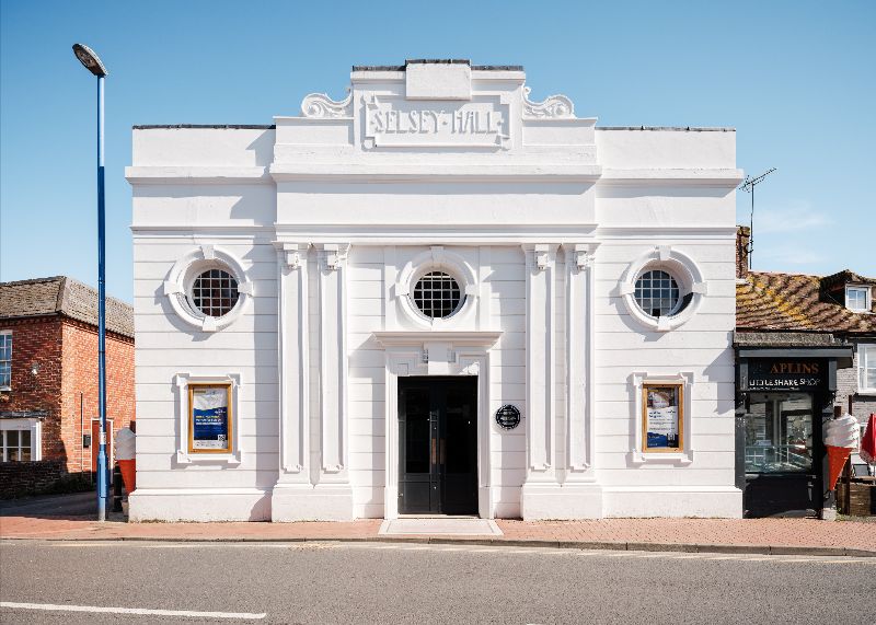 Front view of the restored white art deco Selsey Pavilion building on a sunny day, featuring circular windows, decorative columns, poster display cases, and a central entrance along the high street.