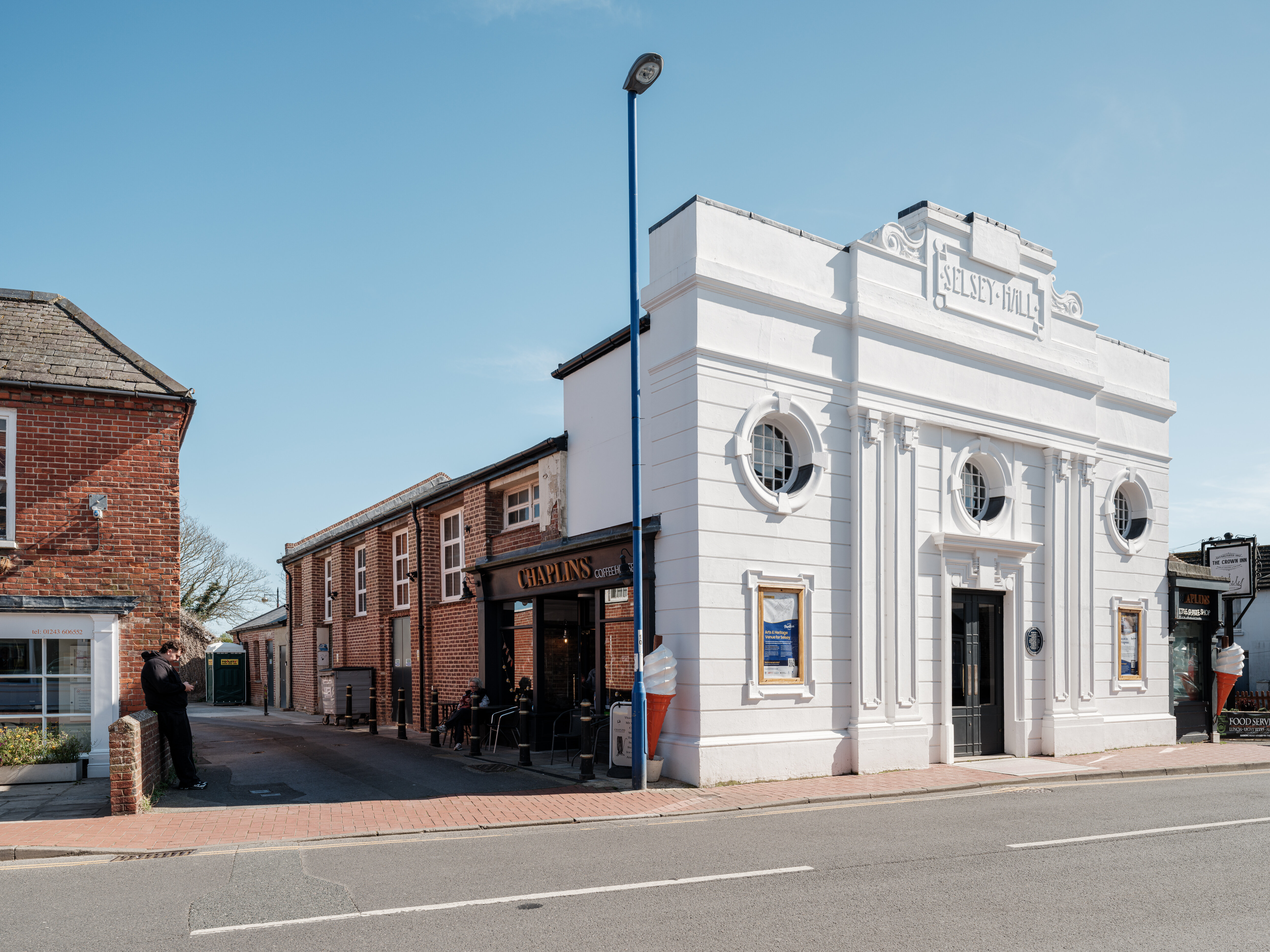 Angled view of the restored white art deco Selsey Hall building on a sunny day, showing its distinctive circular windows, decorative façade, and main entrance. The neighbouring Chaplin’s café and seating area are visible beside the building, with a clear blue sky overhead.