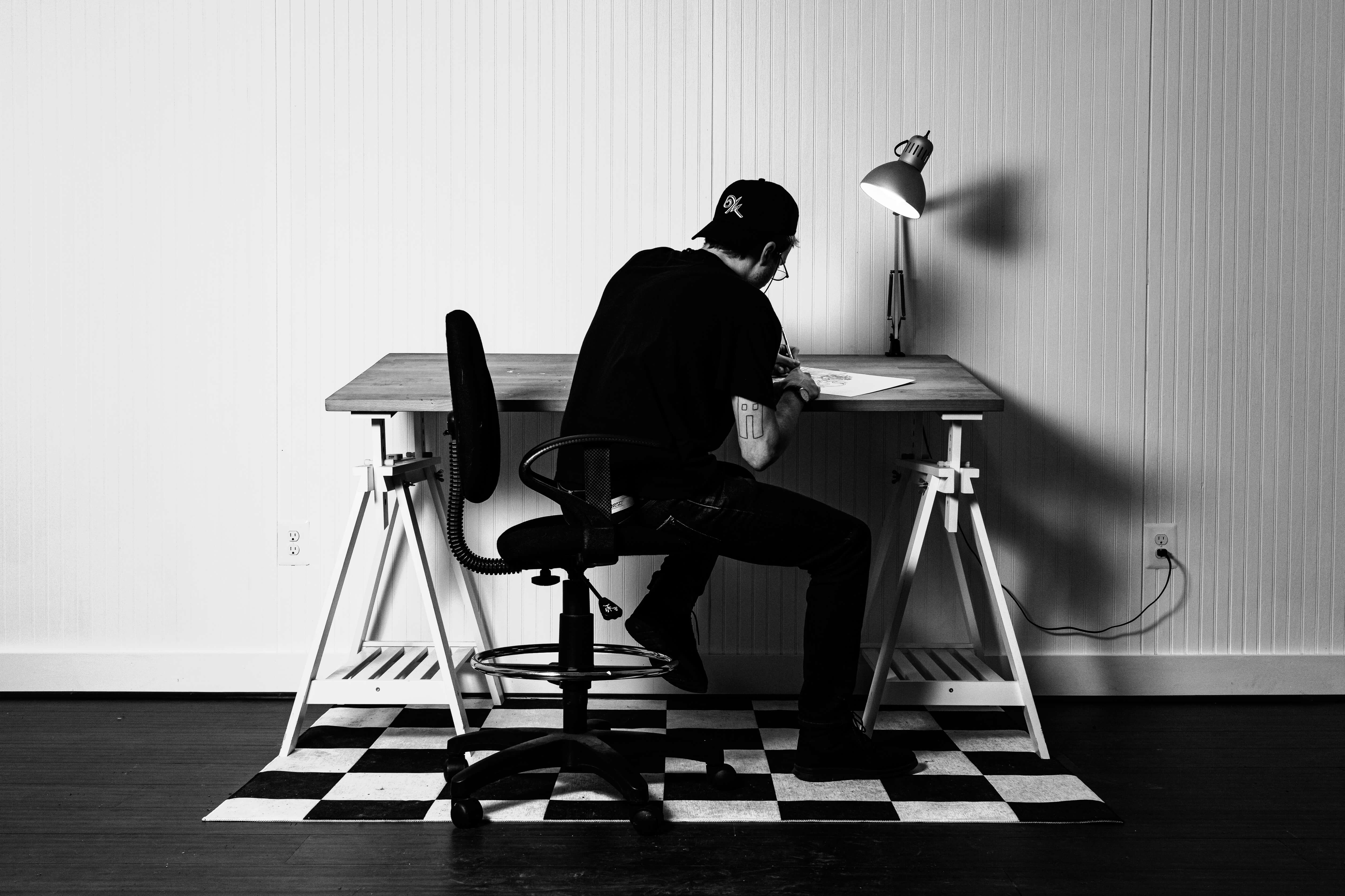 Individual wearing a black cap and shirt sitting on an office chair, drawing at a wooden desk with a lamp in a minimalist room.